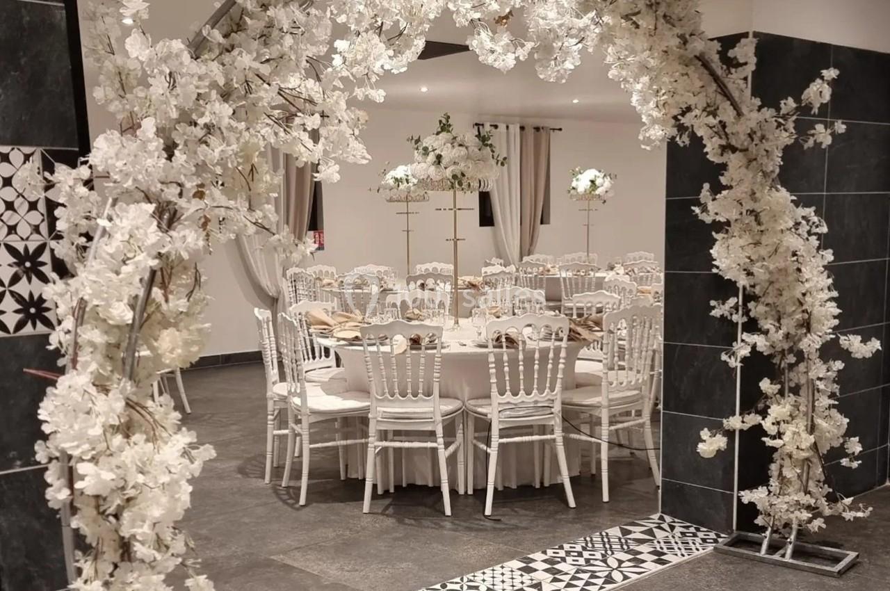 Salle de réception décorée avec des tables rondes, des chaises blanches et une arche florale blanche en premier plan.