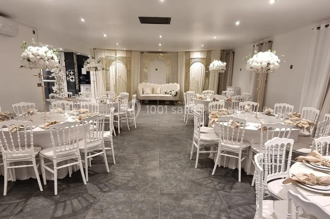 Salle de réception décorée avec des tables rondes, nappes blanches, chaises élégantes et centres de table floraux.