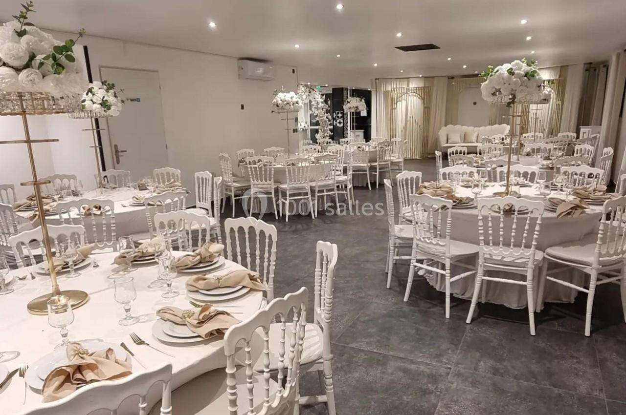 Salle de réception élégamment décorée avec des tables rondes, nappes blanches, chaises blanches et centres de table fleuris.