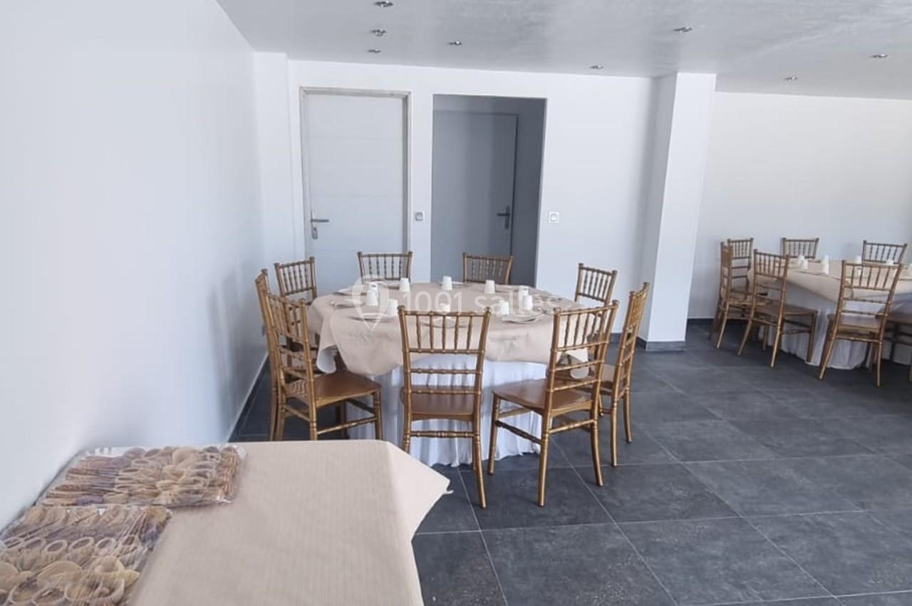 Salle avec tables rondes dressées de nappes blanches, chaises en bois doré et un buffet couvert sur le côté.