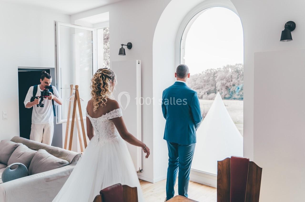 Une mariée s'approche d'un homme en costume bleu, dos tourné, dans une pièce lumineuse avec un photographe.