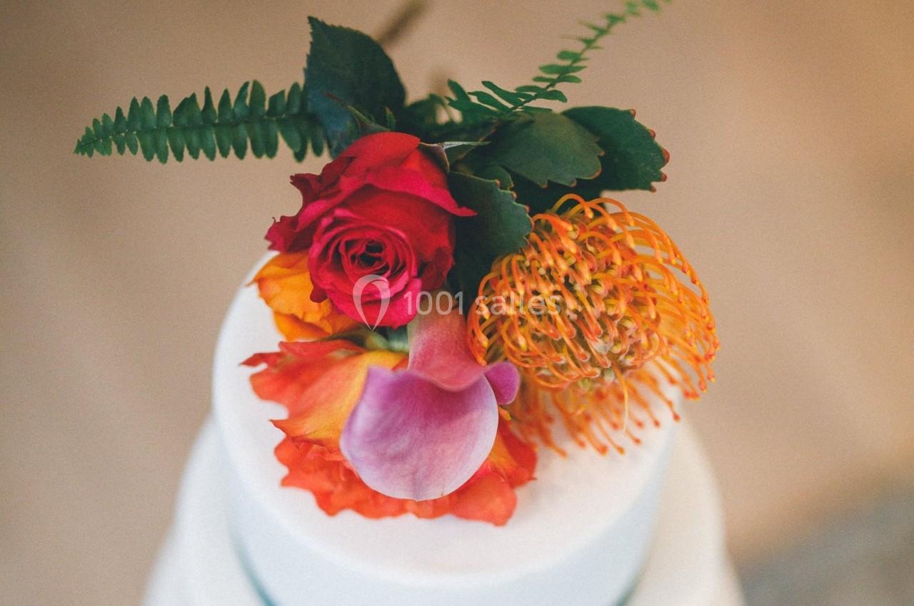 Arrangement floral coloré avec des roses rouges, une fleur orange et des pétales violets sur un gâteau blanc.