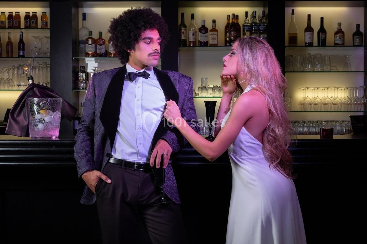 Un homme en costume et une femme en robe élégante interagissent devant un bar éclairé.