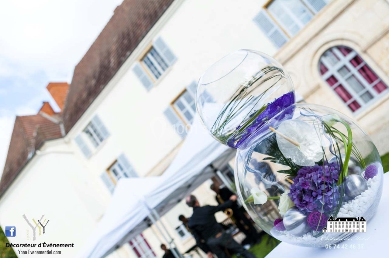 Location salle Roanne (Loire) - Château de Matel #30 Composition florale violette dans un vase en verre devant une grande bâtisse avec des tentes blanches en arrière-plan.