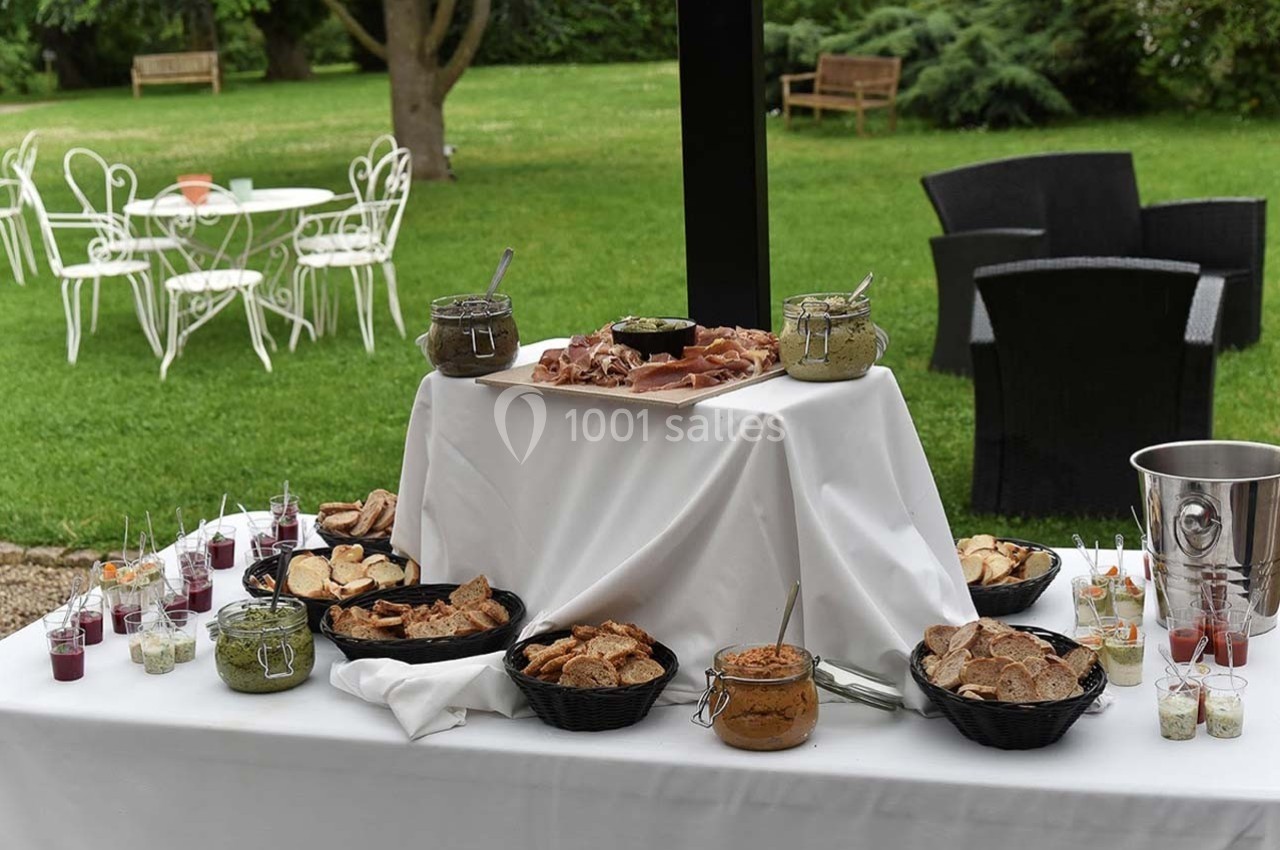 Location salle Roanne (Loire) - Château de Matel #31 Table de buffet en extérieur avec pain, pâtés, condiments et verrines, dans un jardin avec mobilier de jardin en arrière…