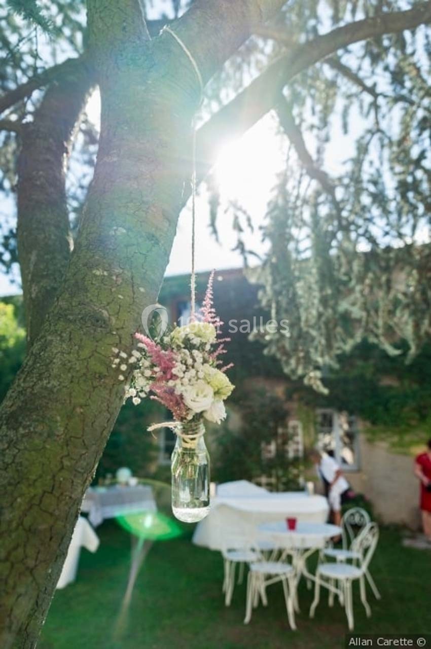 Location salle Roanne (Loire) - Château de Matel #39 Petit vase en verre suspendu à une branche d'arbre, contenant des fleurs, dans un jardin éclairé par le soleil.