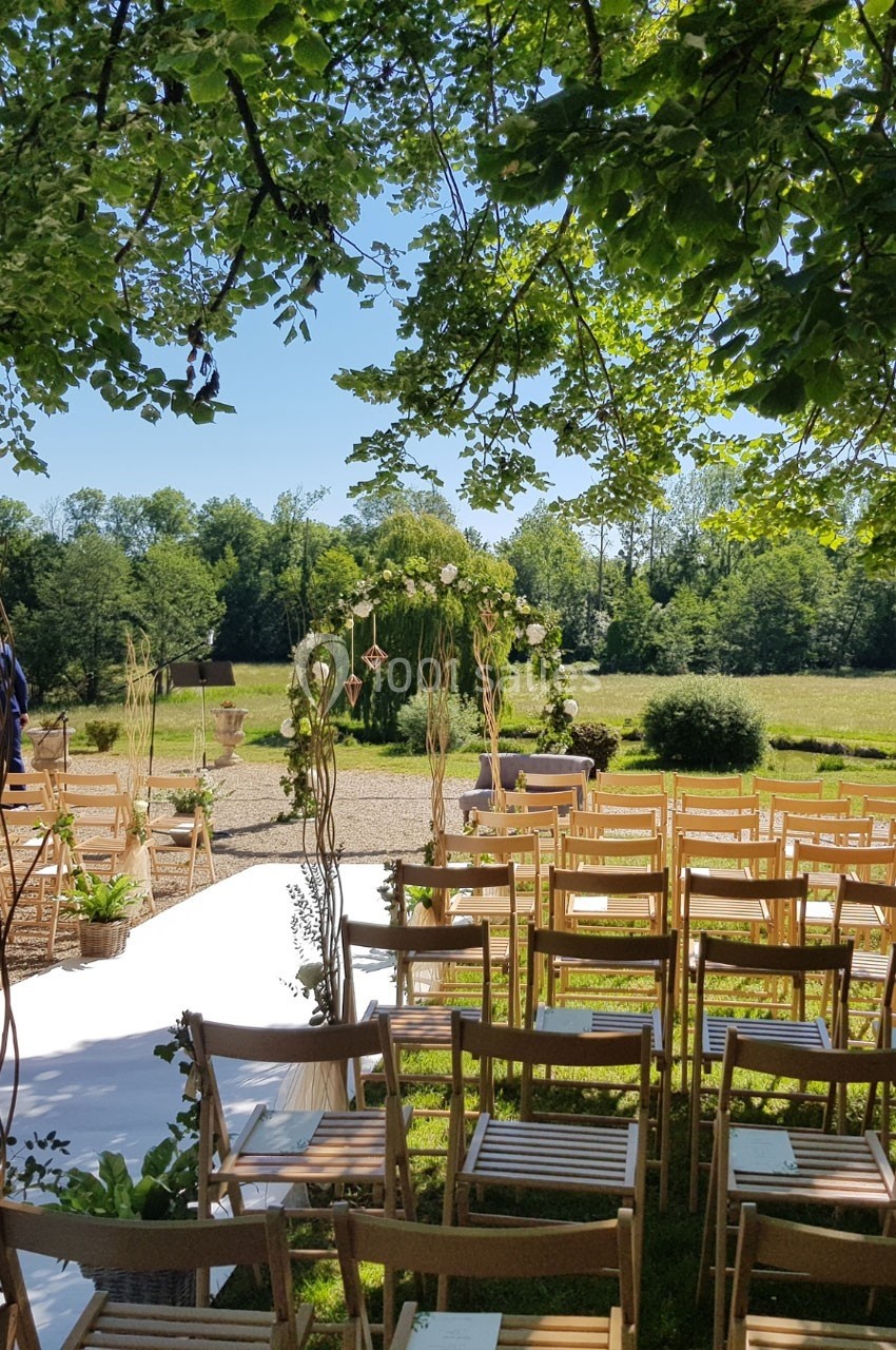 Location salle Roanne (Loire) - Château de Matel #23 Chaises en bois disposées en extérieur sous des arbres, face à une arche décorée pour une cérémonie champêtre.
