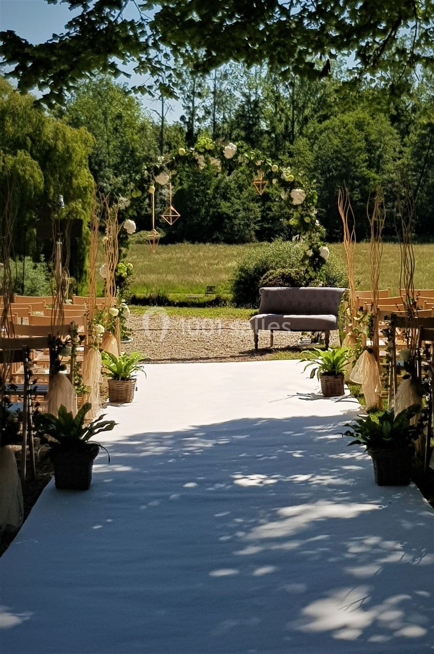 Location salle Roanne (Loire) - Château de Matel #25 Allée décorée pour une cérémonie en plein air, bordée de plantes et menant à une arche florale devant un champ.