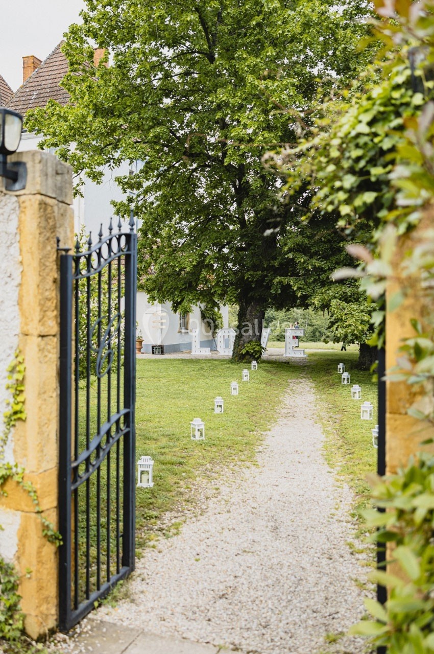Location salle Roanne (Loire) - Château de Matel #45 Allée gravillonnée bordée de lanternes blanches menant à un jardin arboré, vue depuis un portail en fer forgé.