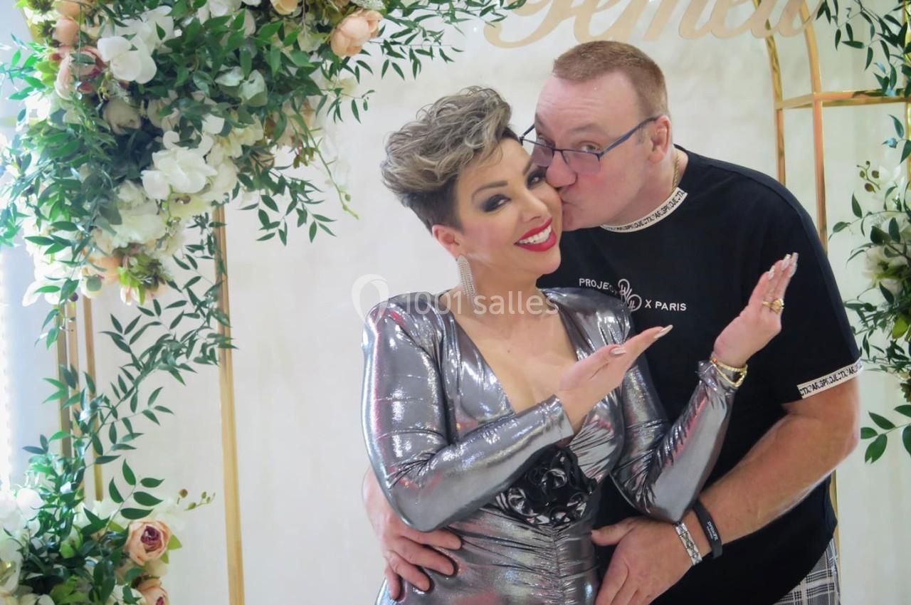 Un homme embrasse une femme souriante vêtue d'une robe argentée, devant une arche décorée de fleurs.