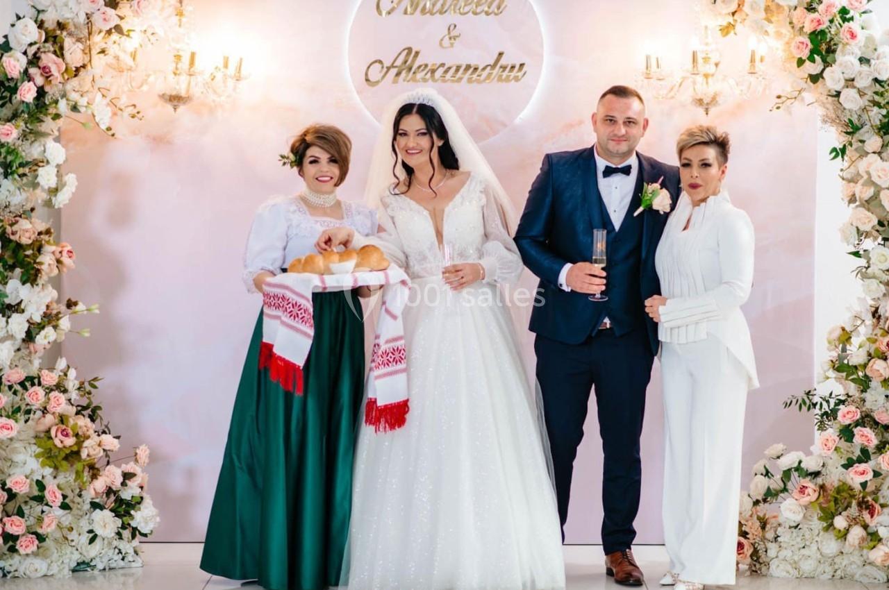 Un couple de mariés pose avec deux invités, entourés de décorations florales et d'un panneau avec leurs prénoms.