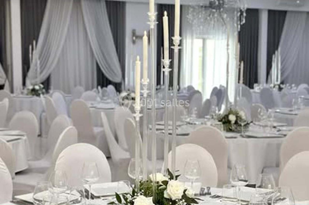 Salle de réception élégante avec tables dressées, nappes blanches, chandeliers et décor floral blanc.