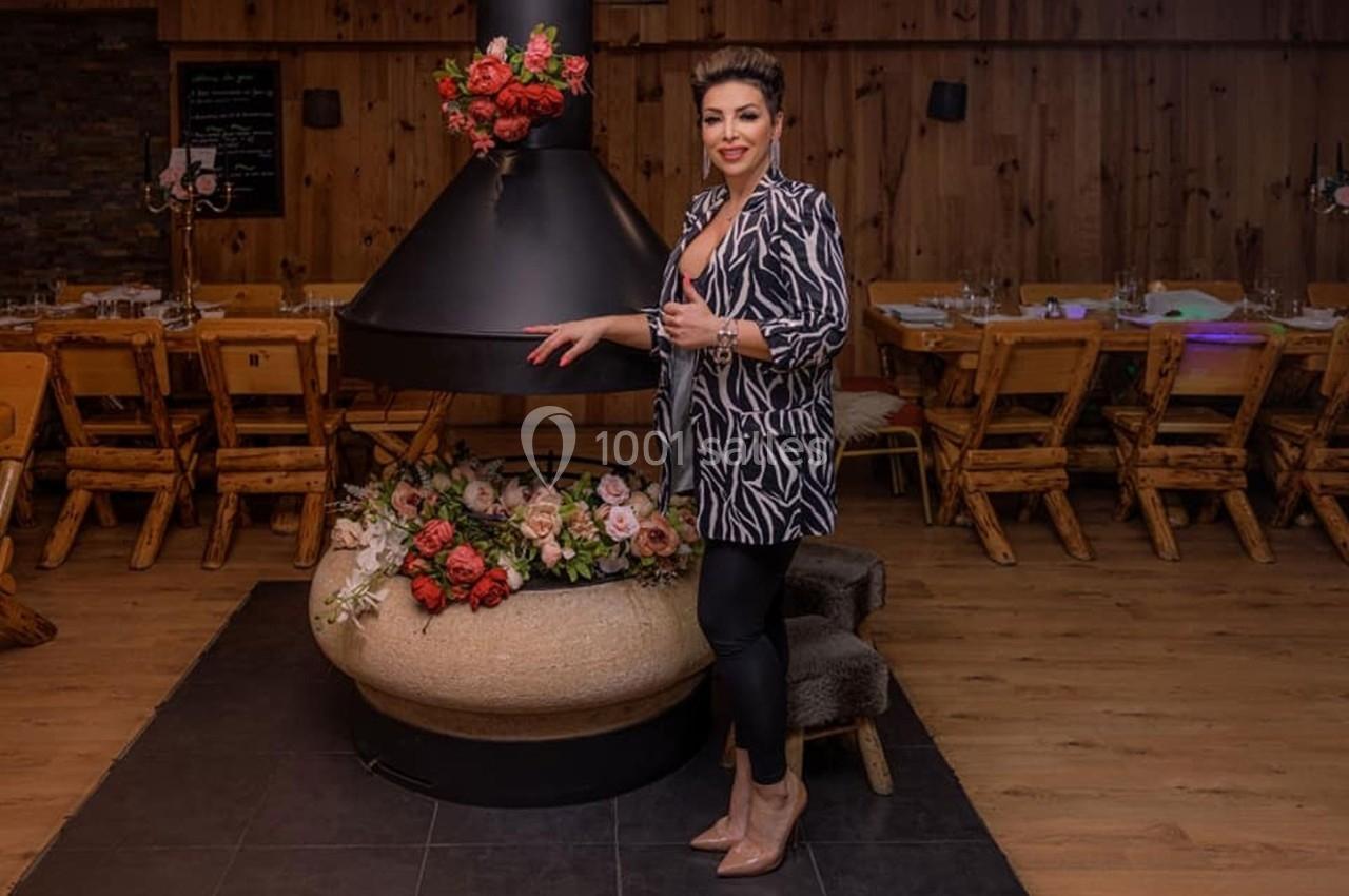 Une femme debout près d'une cheminée décorée de fleurs, dans une salle en bois avec des tables et des chaises rustiques.