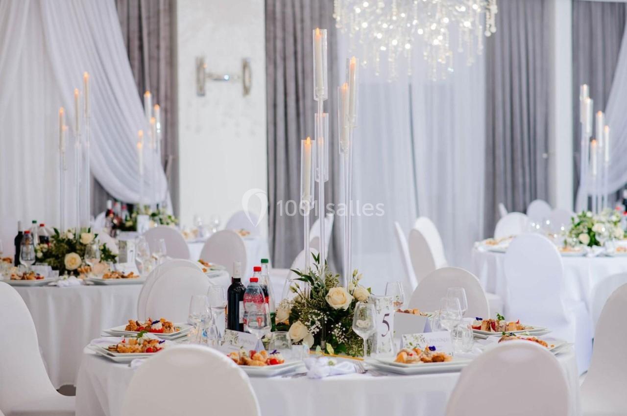 Salle de réception élégante avec tables rondes dressées, décorations florales et chandeliers modernes.