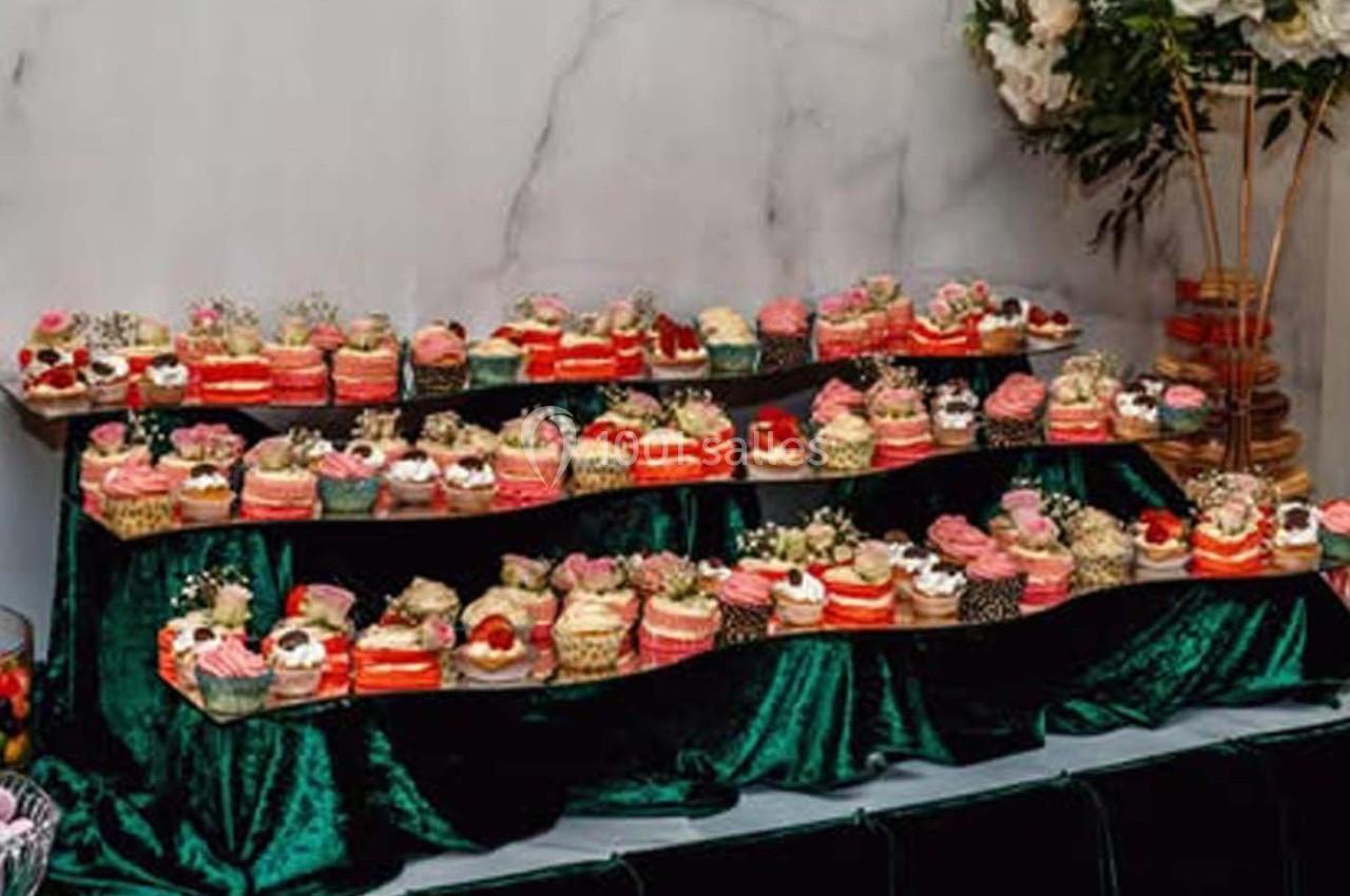 Présentoir de cupcakes variés décorés de fleurs et fruits, disposés sur des étagères recouvertes de tissu vert.