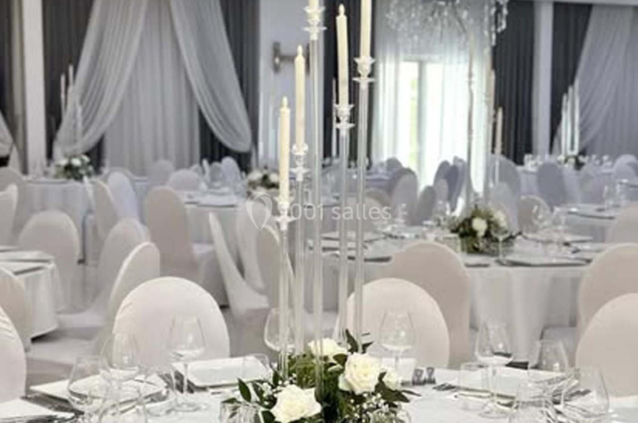 Salle de réception élégante avec tables rondes dressées, nappes blanches, chandeliers et décor floral.