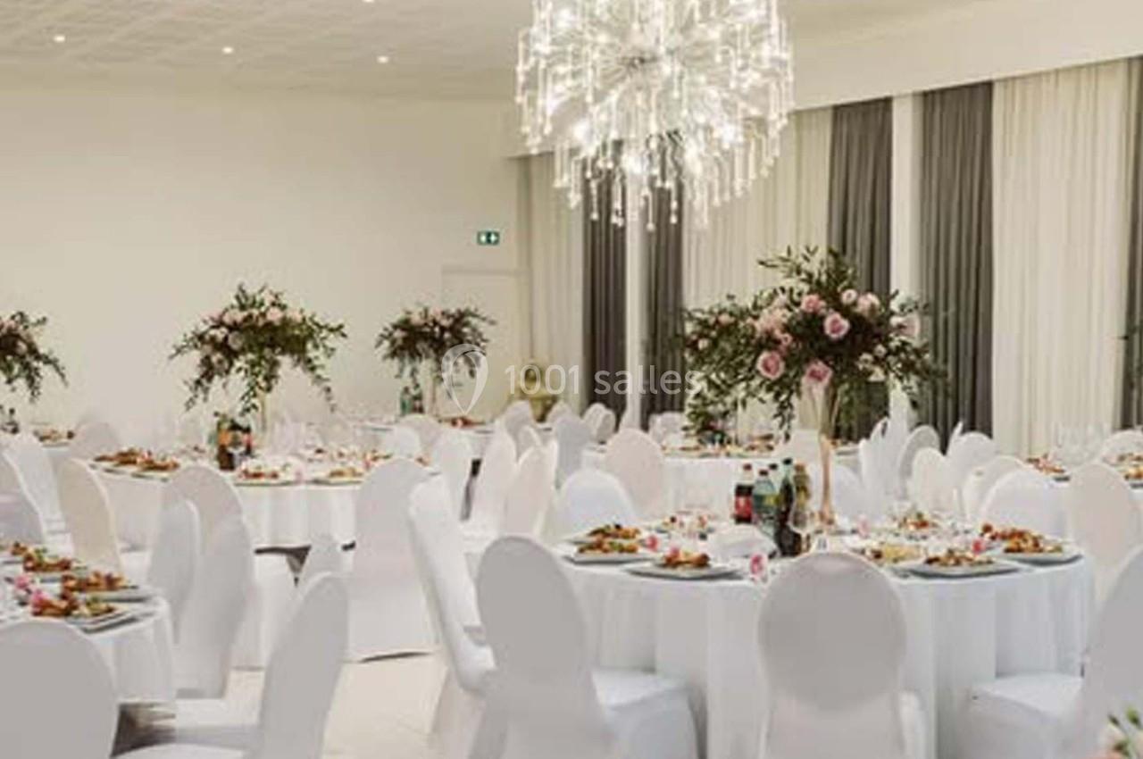 Salle de réception élégante avec tables rondes dressées, chaises blanches et décorations florales.