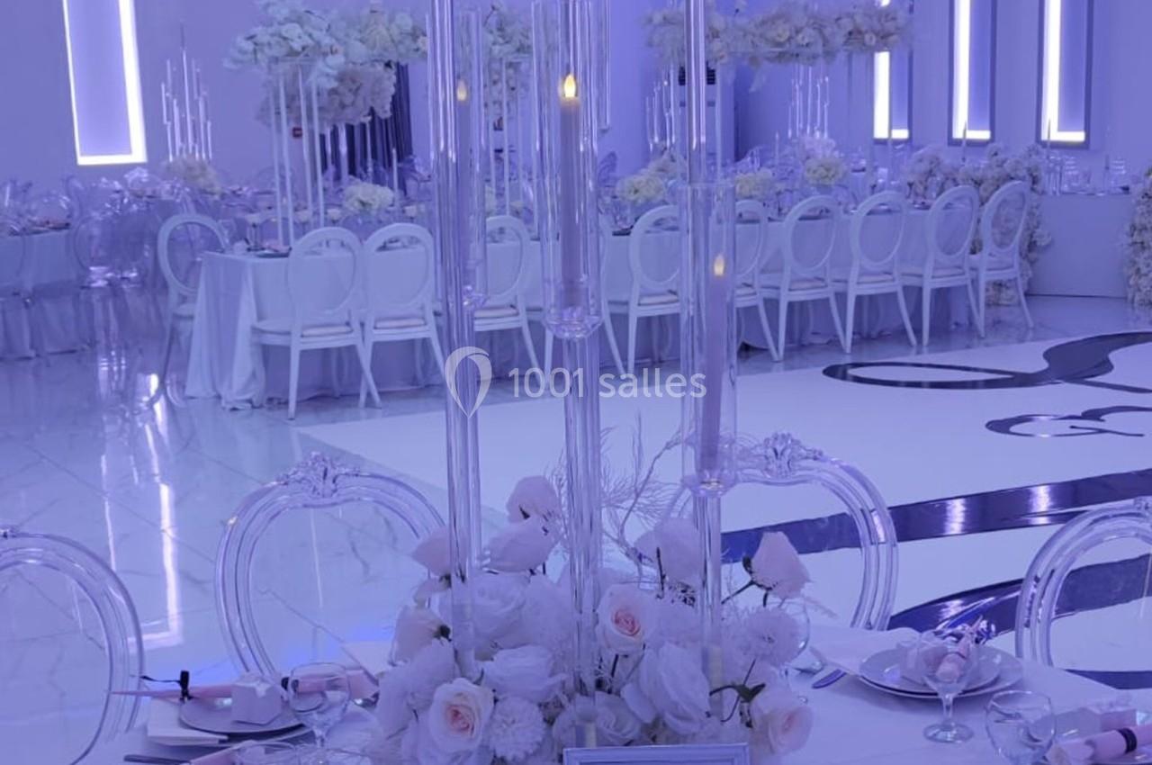 Salle de réception élégante décorée de fleurs blanches, de chandeliers et de tables dressées avec vaisselle raffinée.