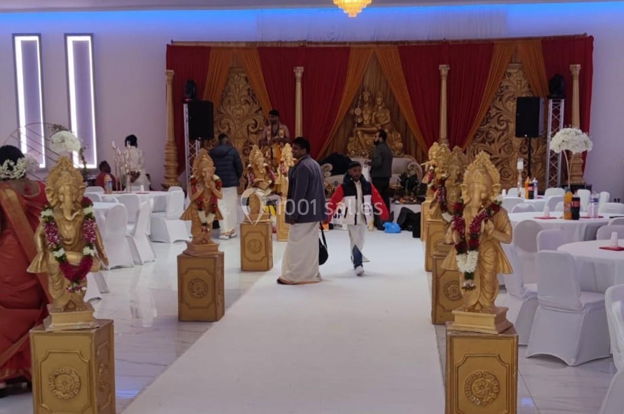 Salle décorée pour un événement avec statues dorées, rideaux rouges et chaises blanches disposées autour de tables.