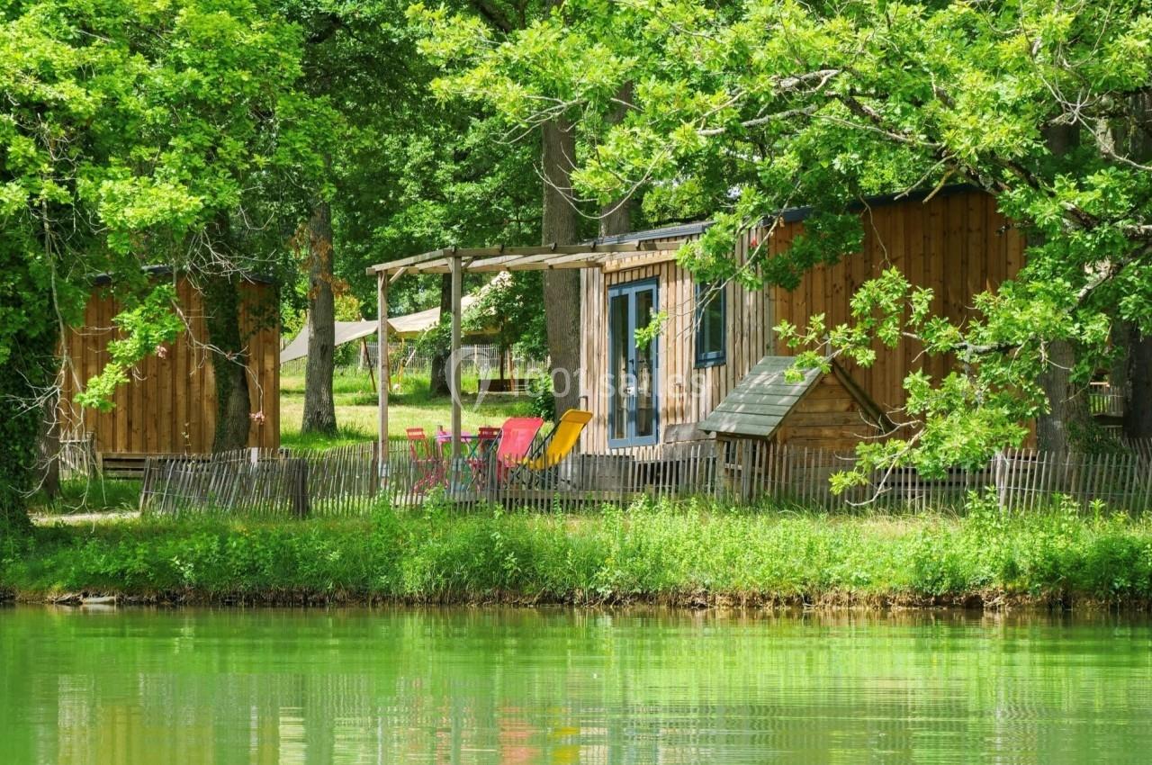 Cabane en bois entourée d'arbres au bord d'un étang, avec des chaises colorées sur une terrasse.