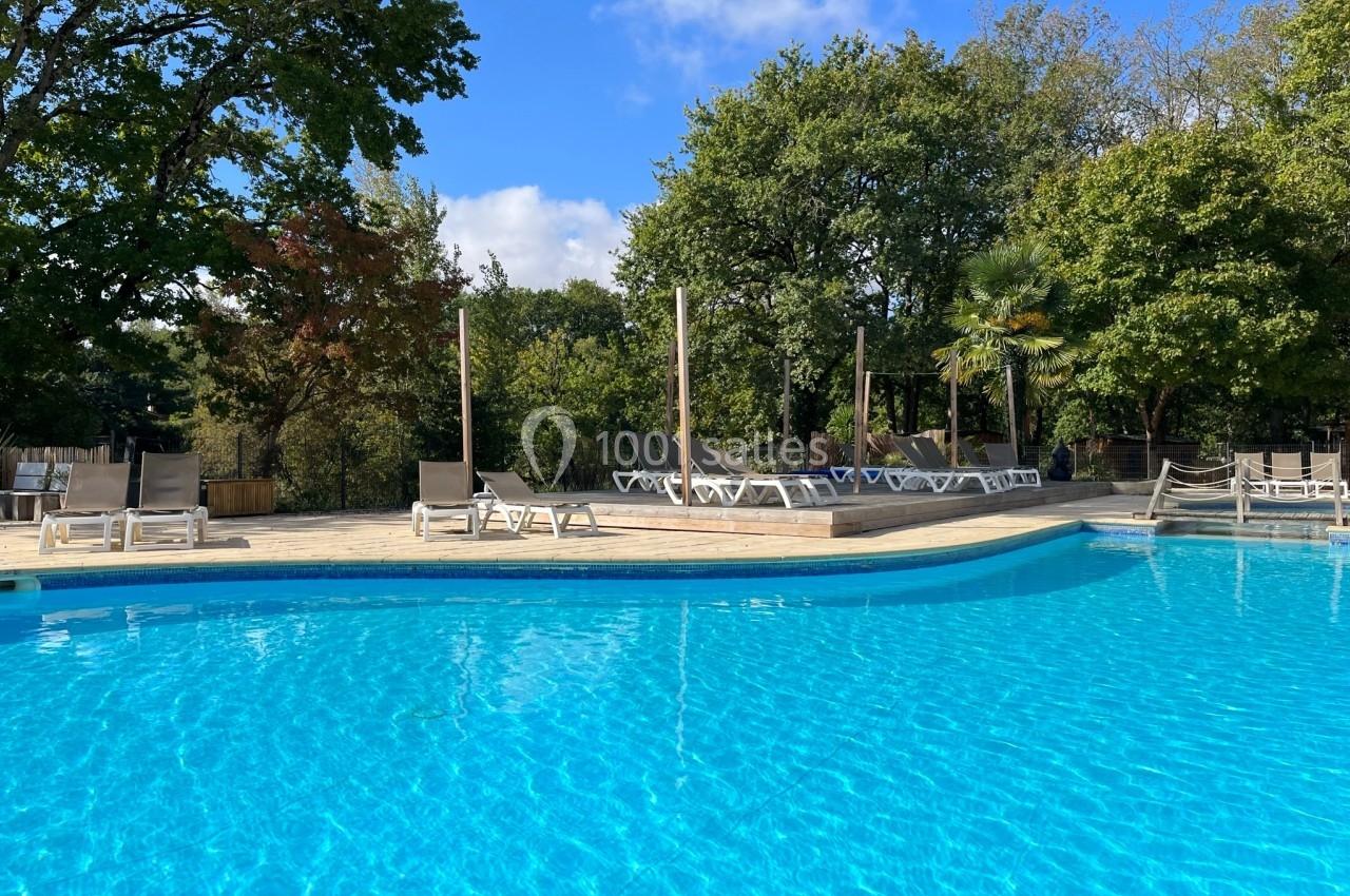 Piscine extérieure entourée de chaises longues et d'arbres sous un ciel bleu clair.