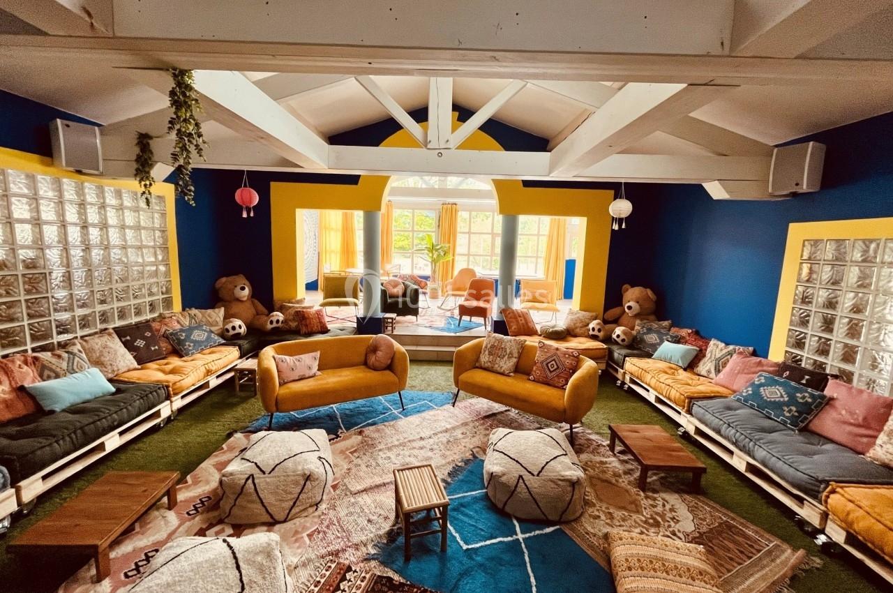 Salle lumineuse avec canapés colorés, coussins, poufs, tapis variés et grandes fenêtres donnant sur un jardin.