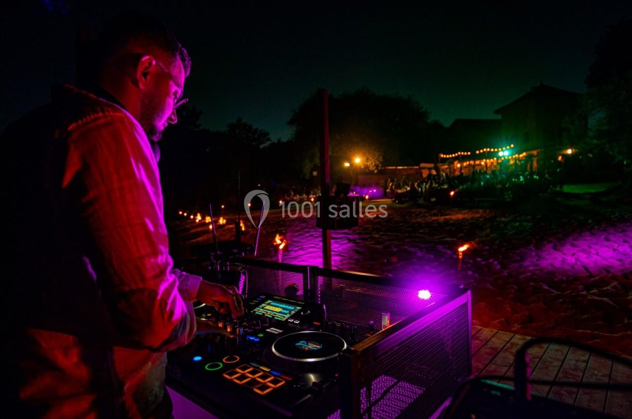 Un DJ mixe de la musique en extérieur la nuit, éclairé par des lumières colorées et entouré d'une ambiance festive.