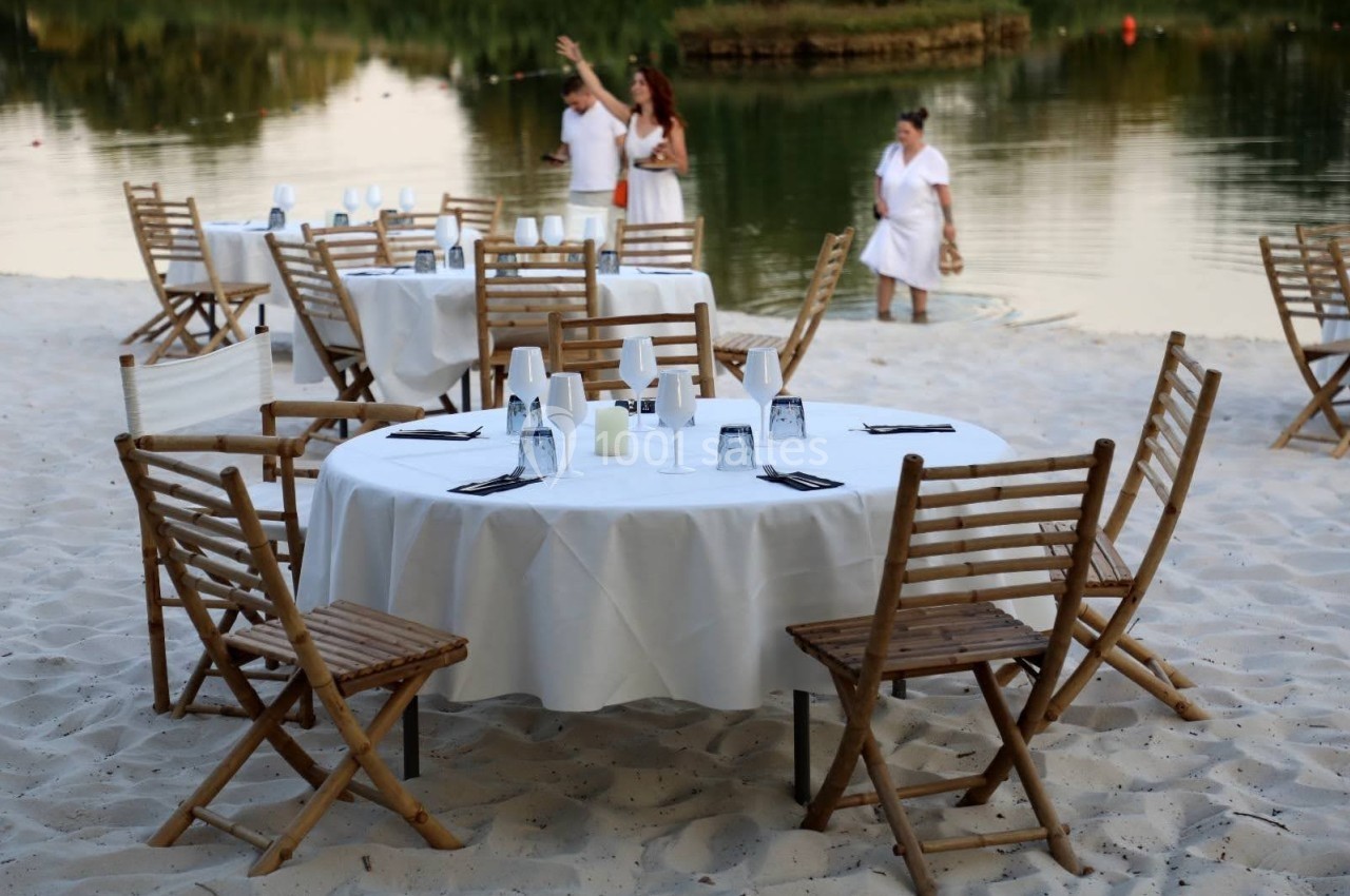 Tables en bois dressées avec nappes blanches sur une plage de sable, avec des personnes près de l'eau en arrière-plan.