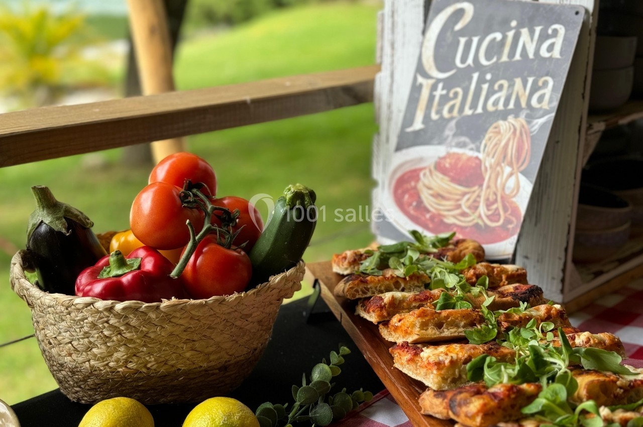 Panier de légumes frais à côté de mini-pizzas garnies, posé sur une table en plein air avec un panneau ’Cucina Italiana’.