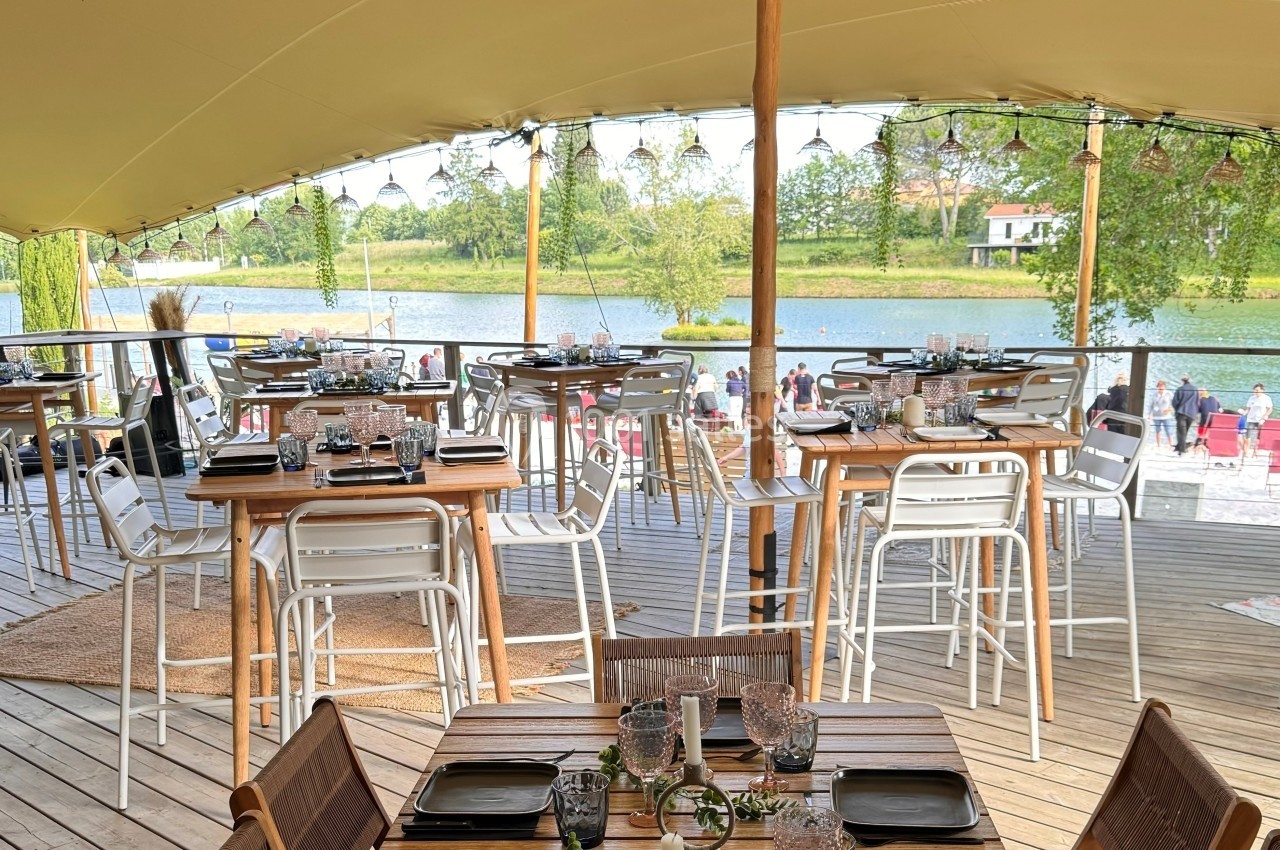 Salle de restaurant en plein air avec tables dressées, vue sur un lac et des arbres en arrière-plan.
