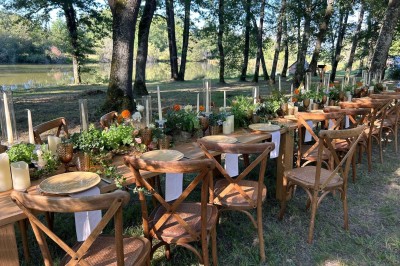 Table en bois décorée de fleurs, bougies et assiettes, installée en extérieur près d'un étang entouré d'arbres.