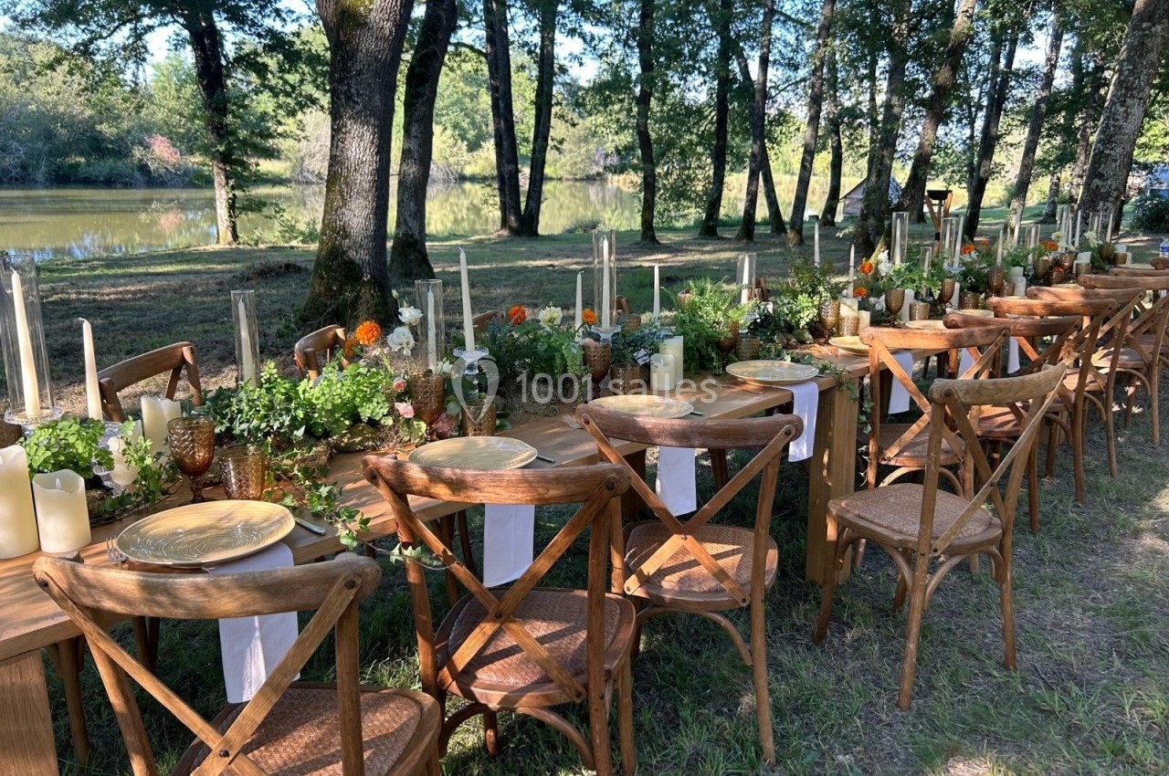 Table en bois décorée de fleurs, bougies et assiettes, installée en extérieur près d'un étang entouré d'arbres.