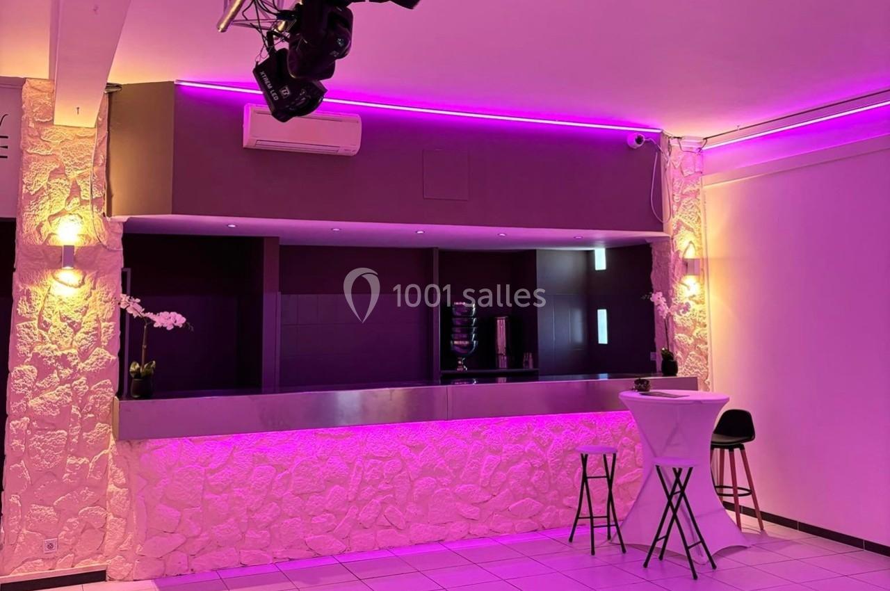 Bar moderne avec comptoir en métal, éclairage violet, murs en pierre blanche et table haute avec tabourets.