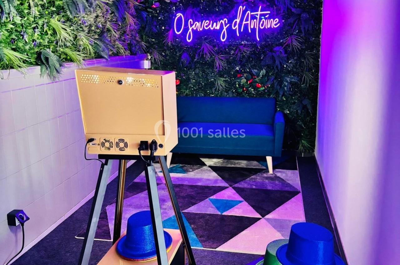 Espace décoré avec un mur végétal, un canapé bleu, un néon violet et un appareil photo sur trépied avec chapeaux colorés.