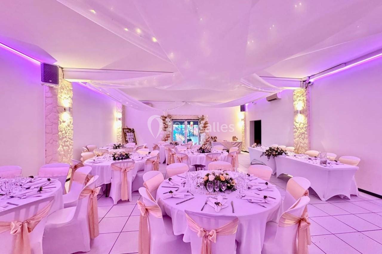 Salle de réception décorée avec des tables rondes, nappes blanches, rubans beige et éclairage violet tamisé.