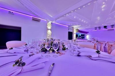 Salle de réception décorée avec des tables rondes, nappes blanches, chaises élégantes et éclairage violet tamisé.