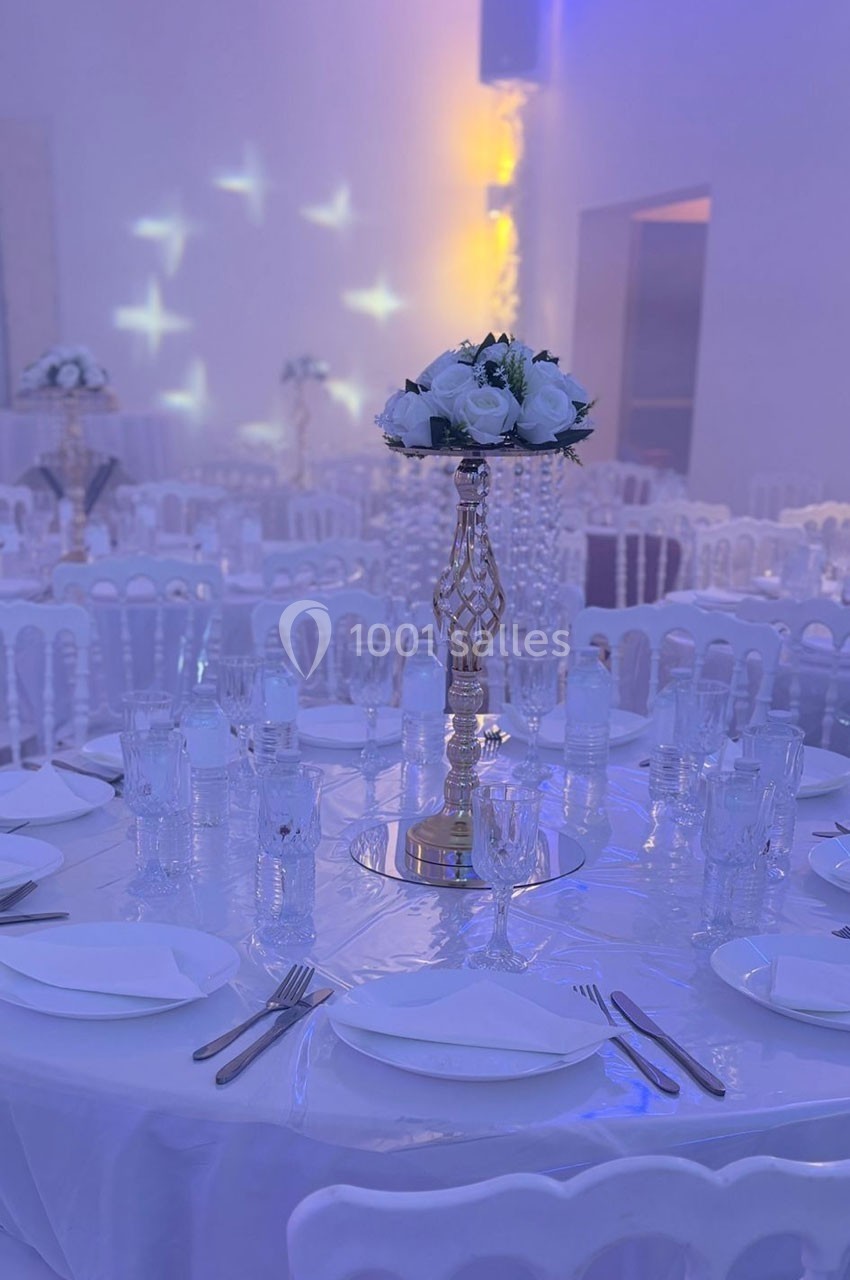 Table de réception élégamment dressée avec nappes blanches, vaisselle, verres et centre de table floral sur support doré.