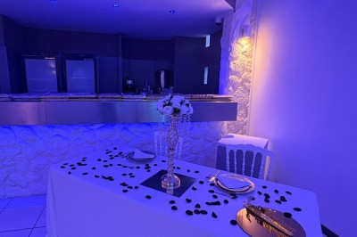 Salle de réception décorée avec des tables rondes, nappes blanches, chaises élégantes et éclairage violet tamisé.