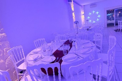 Salle de réception décorée avec des tables rondes, nappes blanches, chaises élégantes et éclairage violet tamisé.
