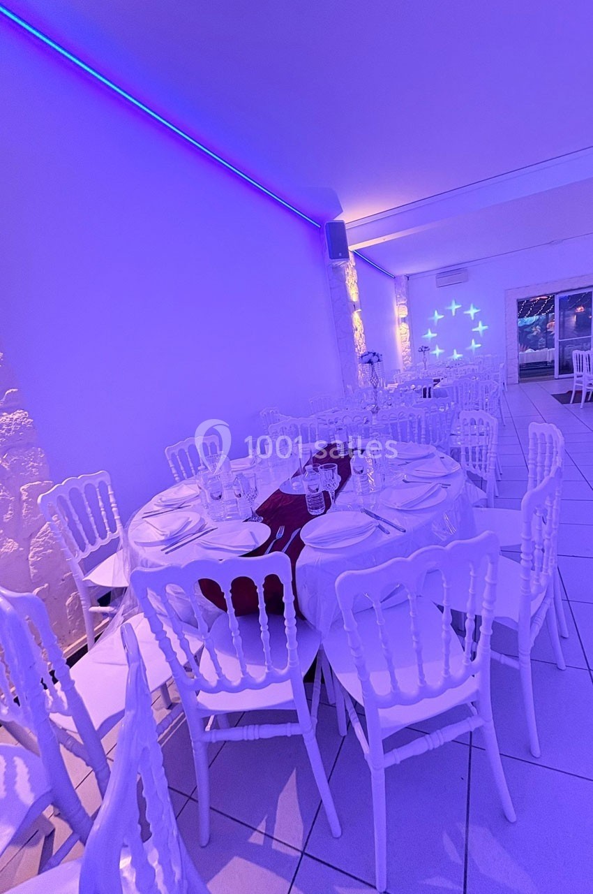 Salle de réception éclairée en violet, avec tables rondes dressées de nappes blanches et chaises assorties.