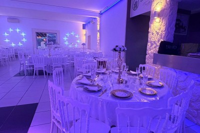 Salle de réception décorée avec des tables rondes, nappes blanches, chaises élégantes et éclairage violet tamisé.