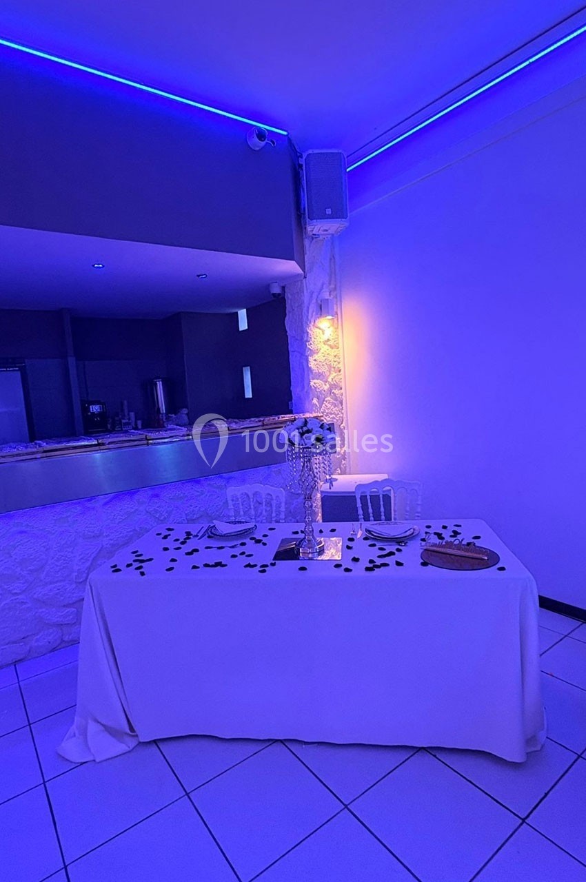 Table décorée avec nappe blanche, chandelier orné de perles, pétales noirs et éclairage bleu en arrière-plan.