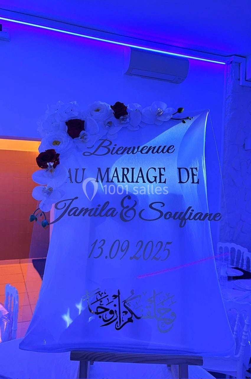 Panneau décoratif avec fleurs blanches et texte ’Bienvenue au mariage de Jamila & Soufiane, 13.09.2025’ sous éclairage bleu.
