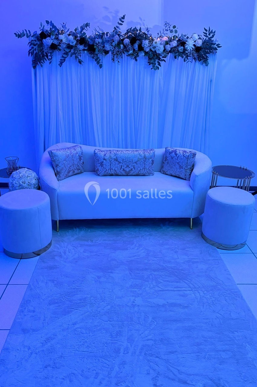 Canapé blanc entouré de poufs, décoré de coussins et fleurs suspendues, éclairé par une lumière bleue.