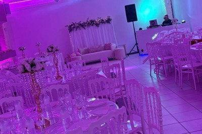 Salle de réception décorée avec des tables rondes, nappes blanches, chaises élégantes et éclairage violet tamisé.