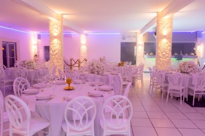 Salle de réception décorée avec des tables rondes, nappes blanches, chaises élégantes et éclairage violet tamisé.