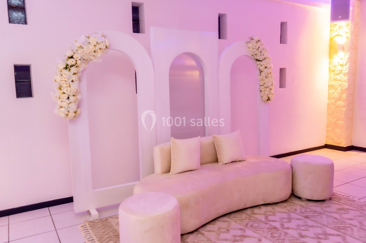 Décor intérieur avec arche blanche ornée de fleurs blanches, canapé beige incurvé et poufs assortis sur un tapis.