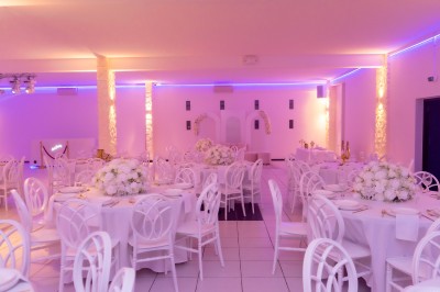 Salle de réception décorée avec des tables rondes, nappes blanches, chaises élégantes et éclairage violet tamisé.