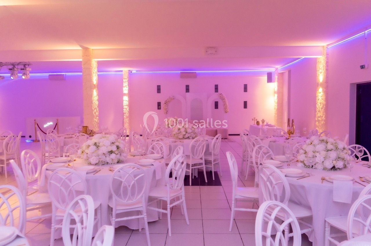 Salle de réception décorée avec des tables rondes, nappes blanches, chaises assorties et éclairage violet tamisé.
