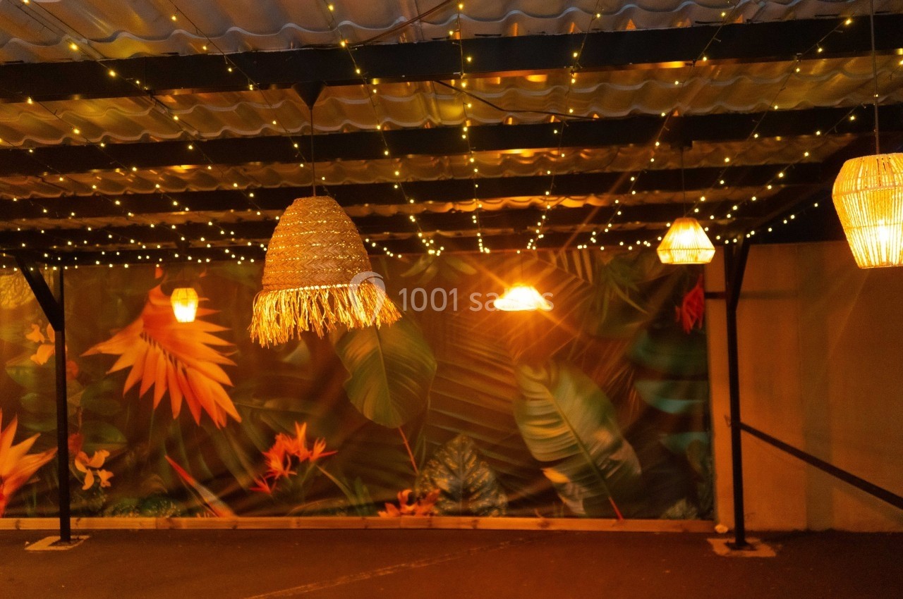 Luminaires suspendus sous une structure métallique, décorés de guirlandes lumineuses et d'un fond tropical coloré.